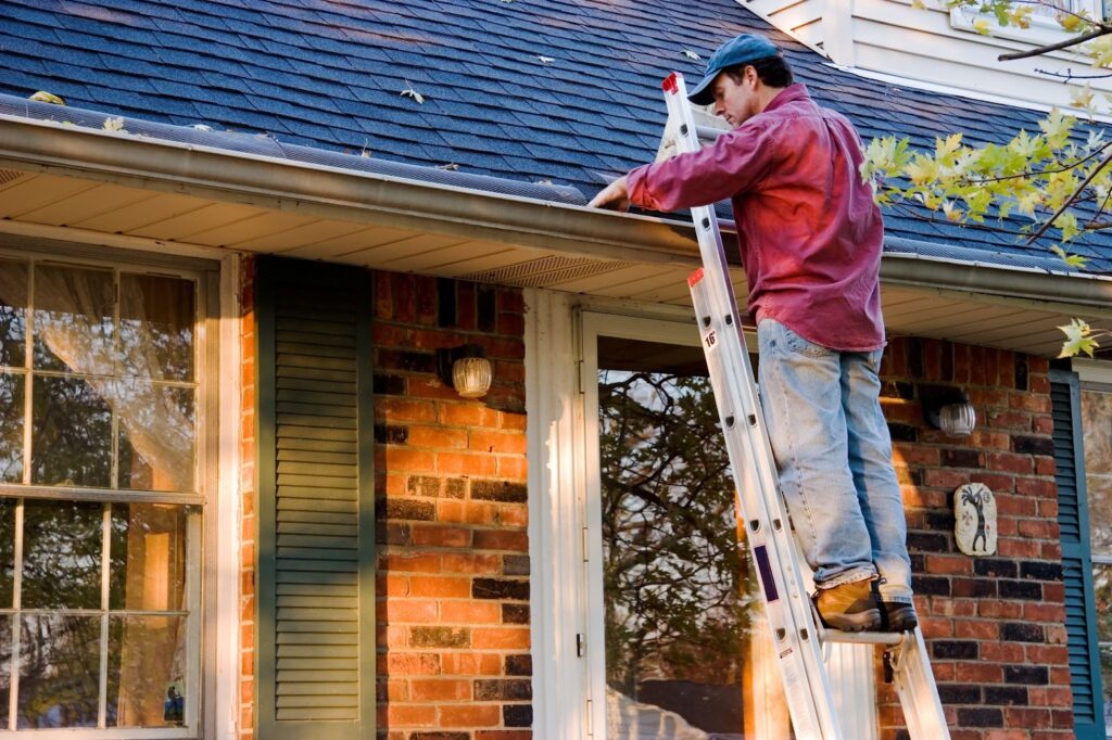A person on a ladder cleans the gutters of a brick house with a blue roof.
