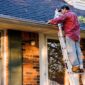 A person on a ladder cleans the gutters of a brick house with a blue roof 85x85