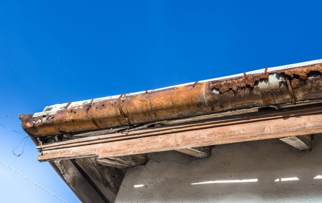 A rusty, weathered gutter on a house roof under a clear blue sky.
