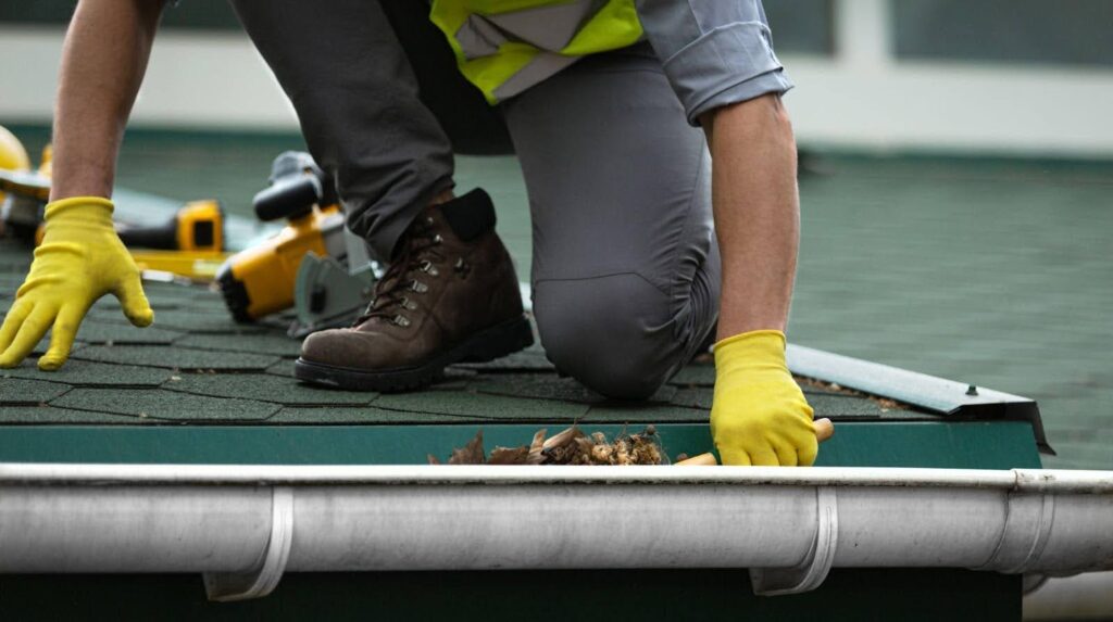 A man wearing yellow gloves cleans a gutter removing debris to ensure proper drainage