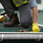 A man wearing yellow gloves cleans a gutter removing debris to ensure proper drainage 85x85
