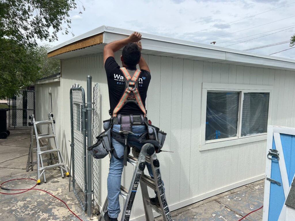 A worker in a safety harness installing a roof edge on a building using a ladder.