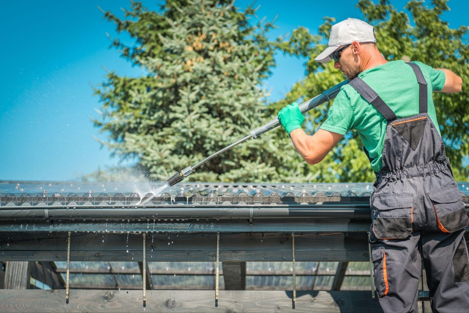 A man in overalls and gloves is cleaning a roof using a brush to remove debris under a clear blue sky