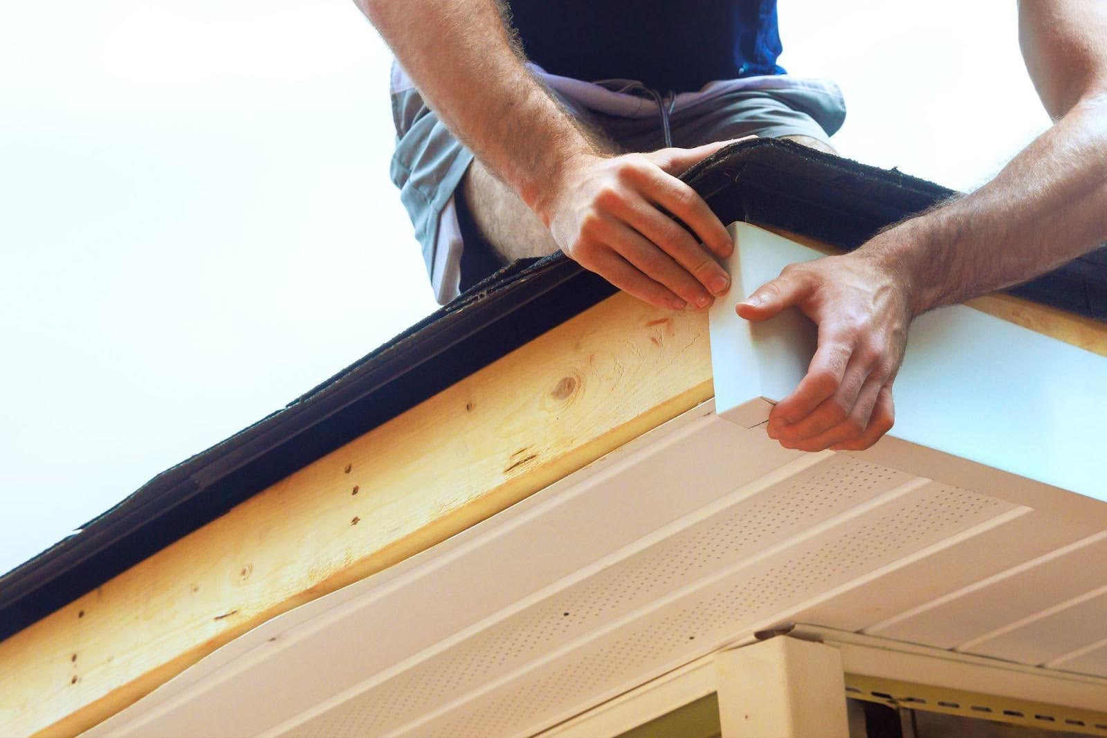 A man works on a roof installing a new gutter with tools in hand and a focused expression on his face