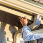 A worker uses a power drill to install or repair a tan metal gutter and downspout system along the roofline of a stucco house 85x85