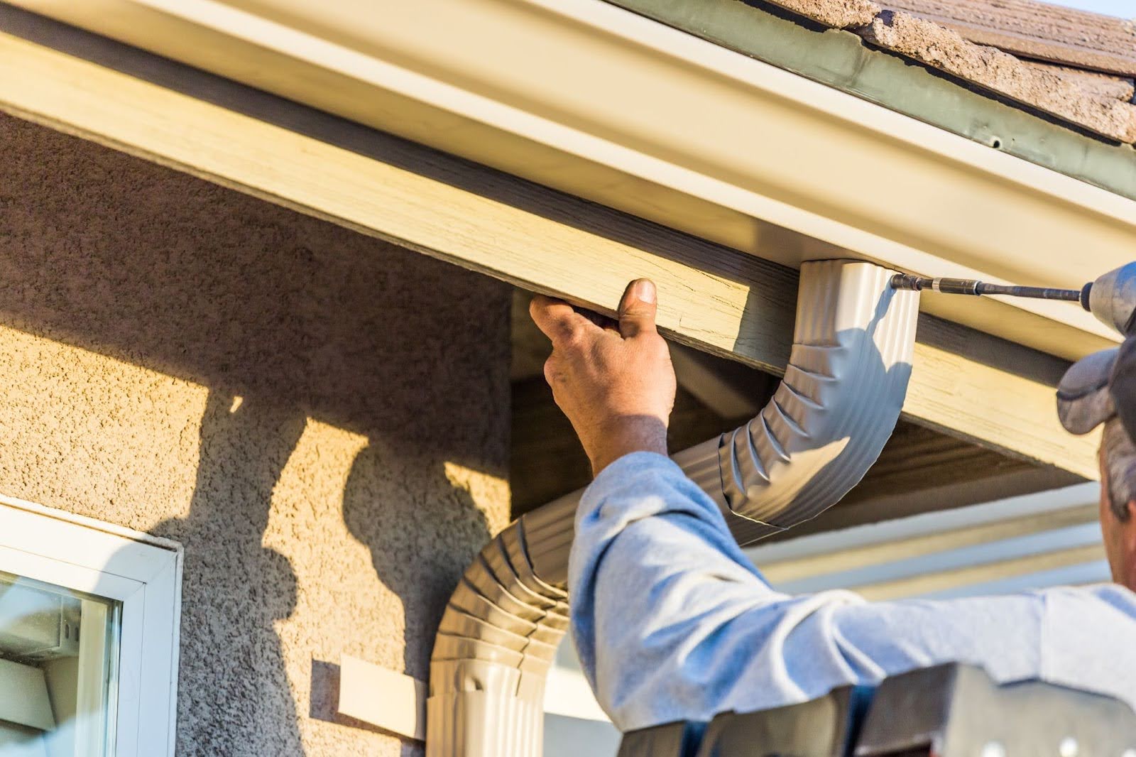 A worker uses a power drill to install or repair a tan metal gutter and downspout system along the roofline of a stucco house.