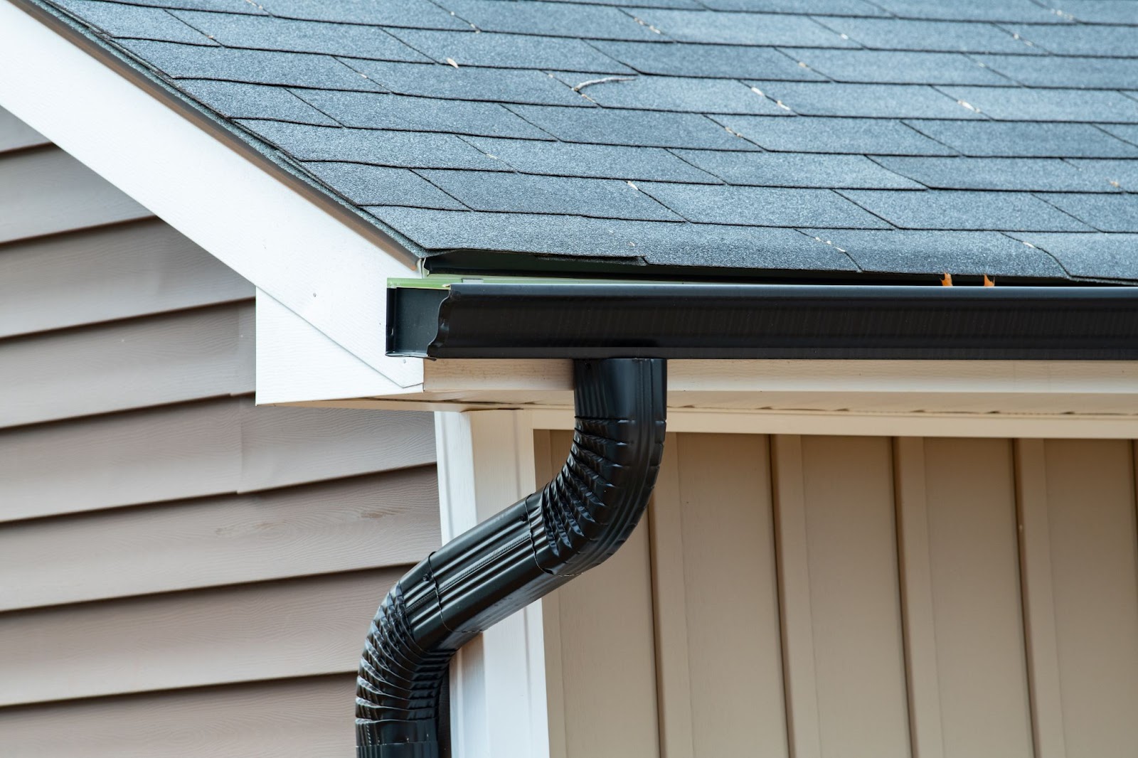 Close-up of a black metal rain gutter and downspout installed on a house with grey shingles and tan siding.